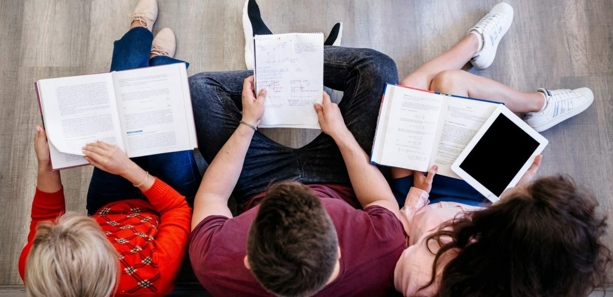 Classificação de sílaba: fotografia tirada de cima de três pessoas estudando, elas estão com cadernos e livros no colo.