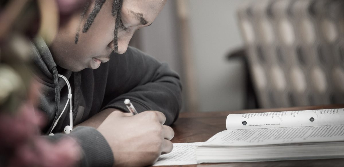 Competências do Enem - fotografia de um adolescente sentado à mesa e escrevendo em uma folha de papel com uma caneta. Ao lado da folha, está um livro aberto.